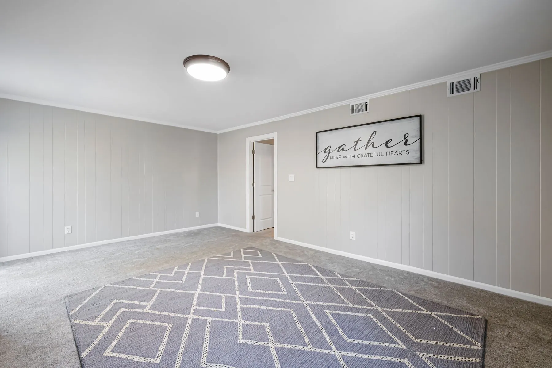An empty finished basement family room with grey wall to wall carpet and a geometric area rug