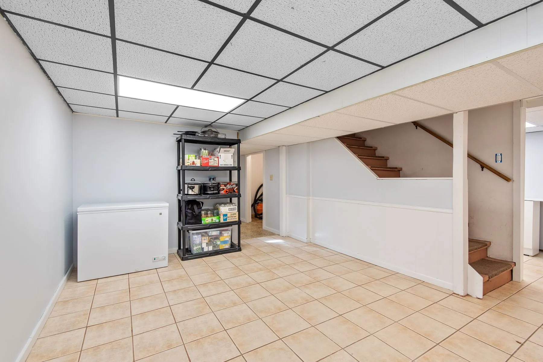 A finished basement with a tiled floor, a drop ceiling, storage shelving, a chest freezer and carpeted stairs