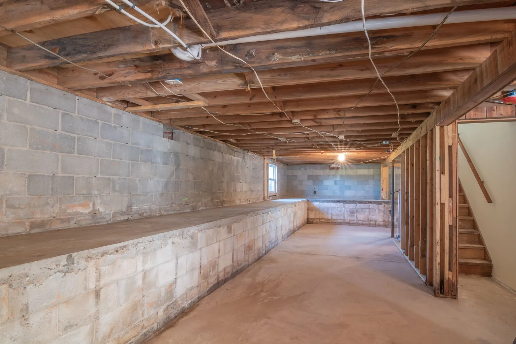 An unfinished basement with concrete block walls, exposed joists and wiring, and a wooden stair
