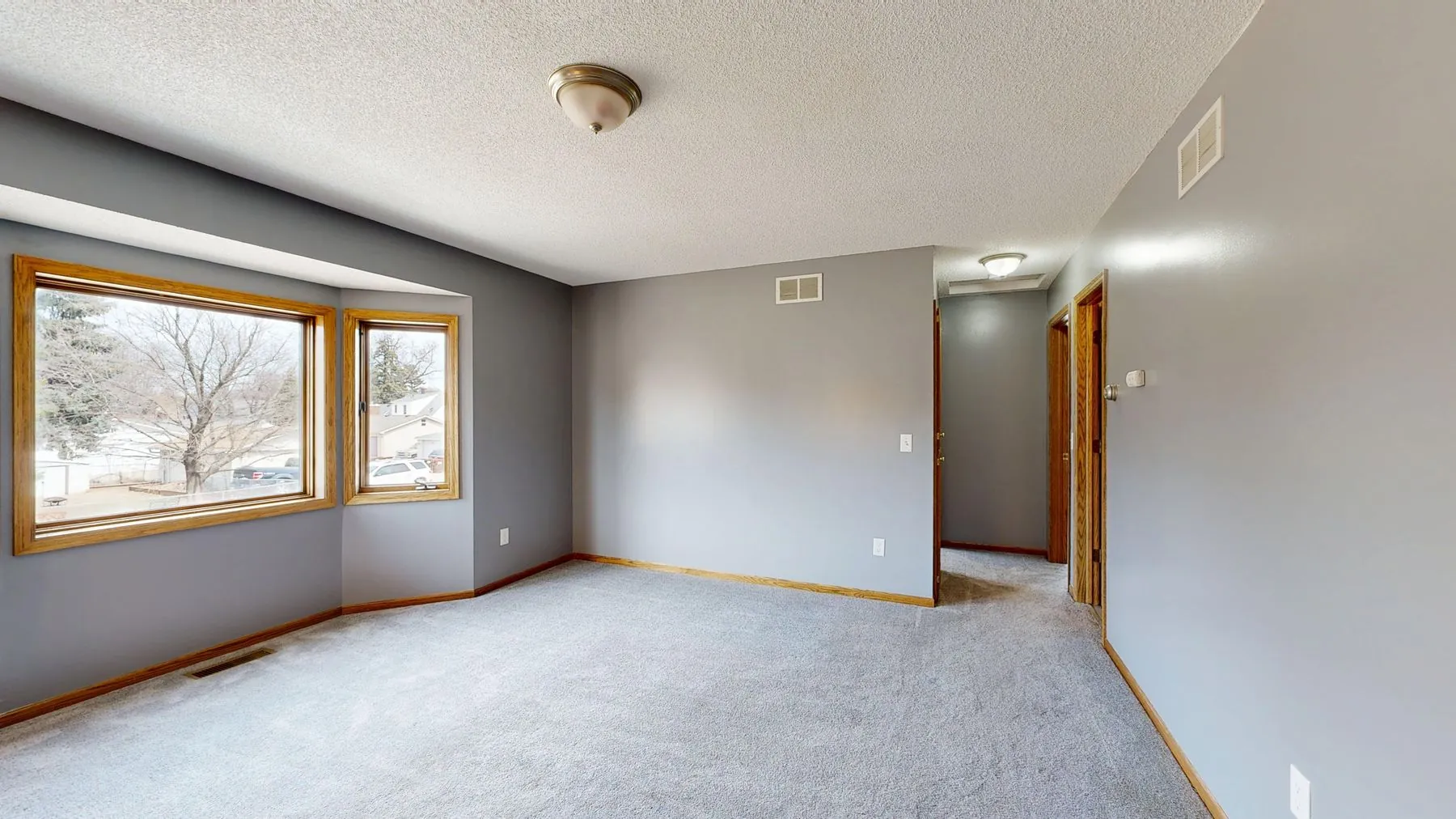 An empty bedroom with grey wall to wall carpet, grey walls, oak trim and a bay window onto a street