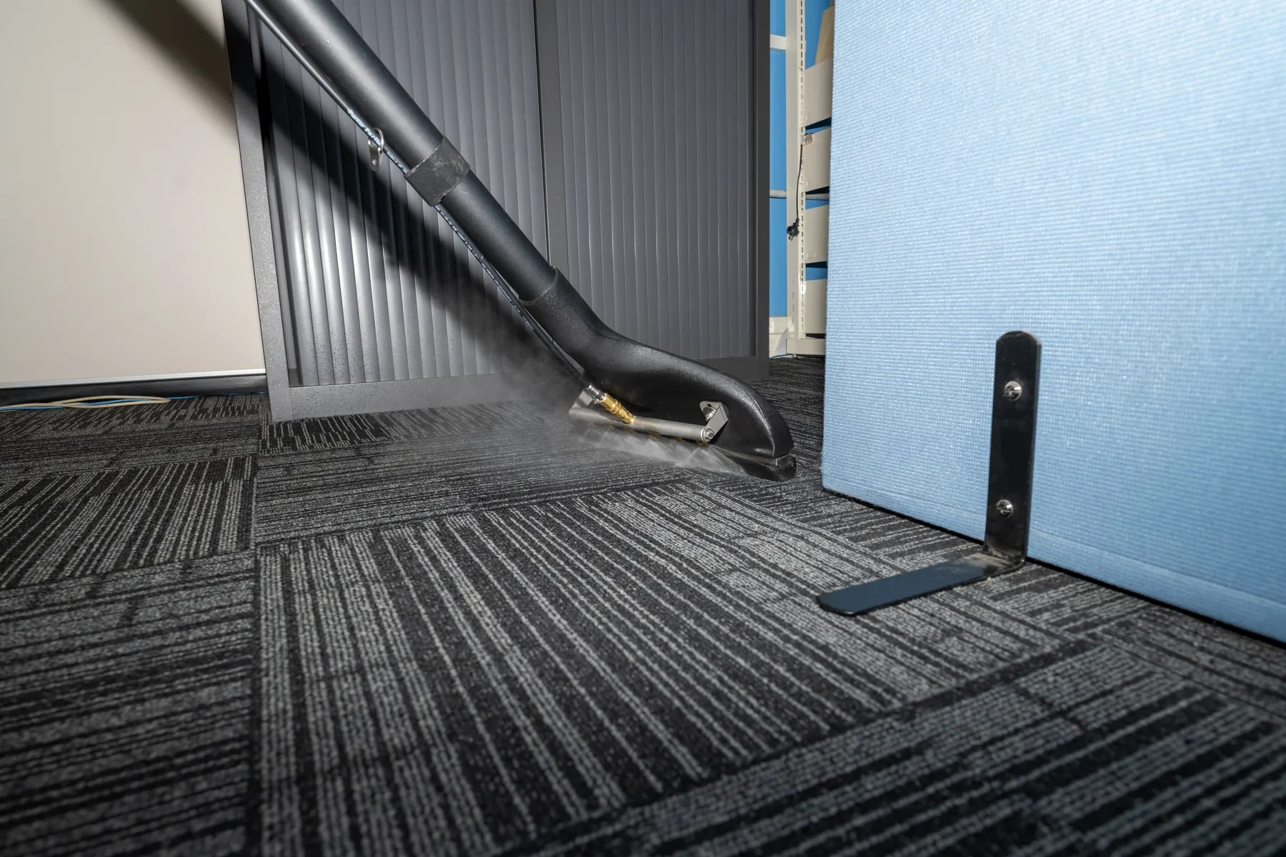 Steam rising from an extraction wand head on charcoal office carpet tiles beside a pale blue acoustic screen