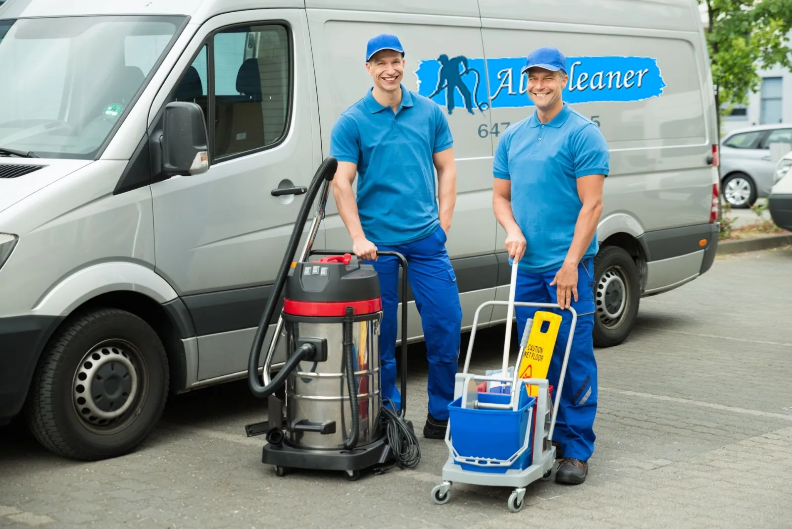 Two AllCleaner technicians in blue uniforms beside the company van, with an extraction vacuum and a mop cart