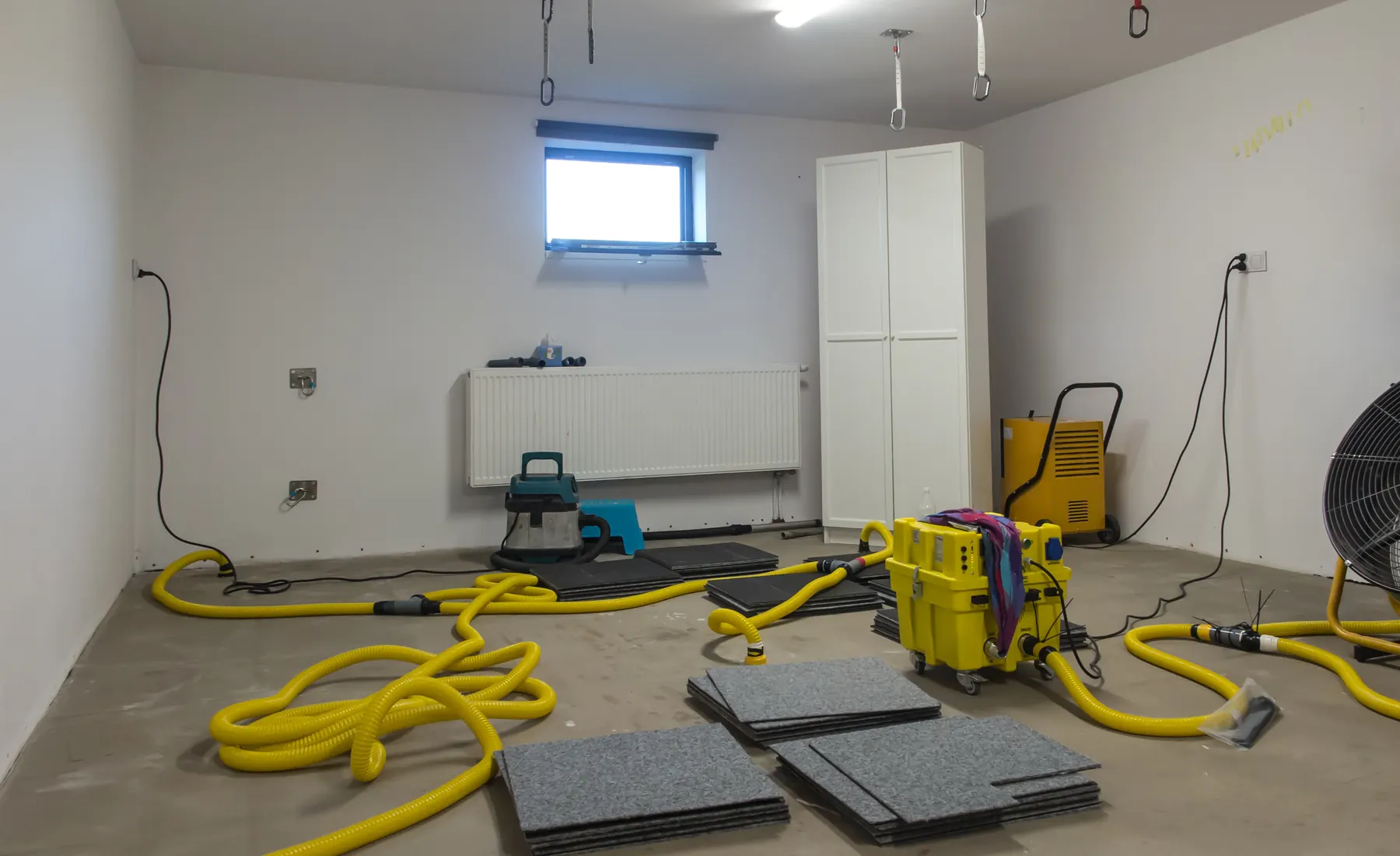 Coiled yellow drying hoses, a drying unit and a drum fan in an empty below grade room with lifted floor boards stacked