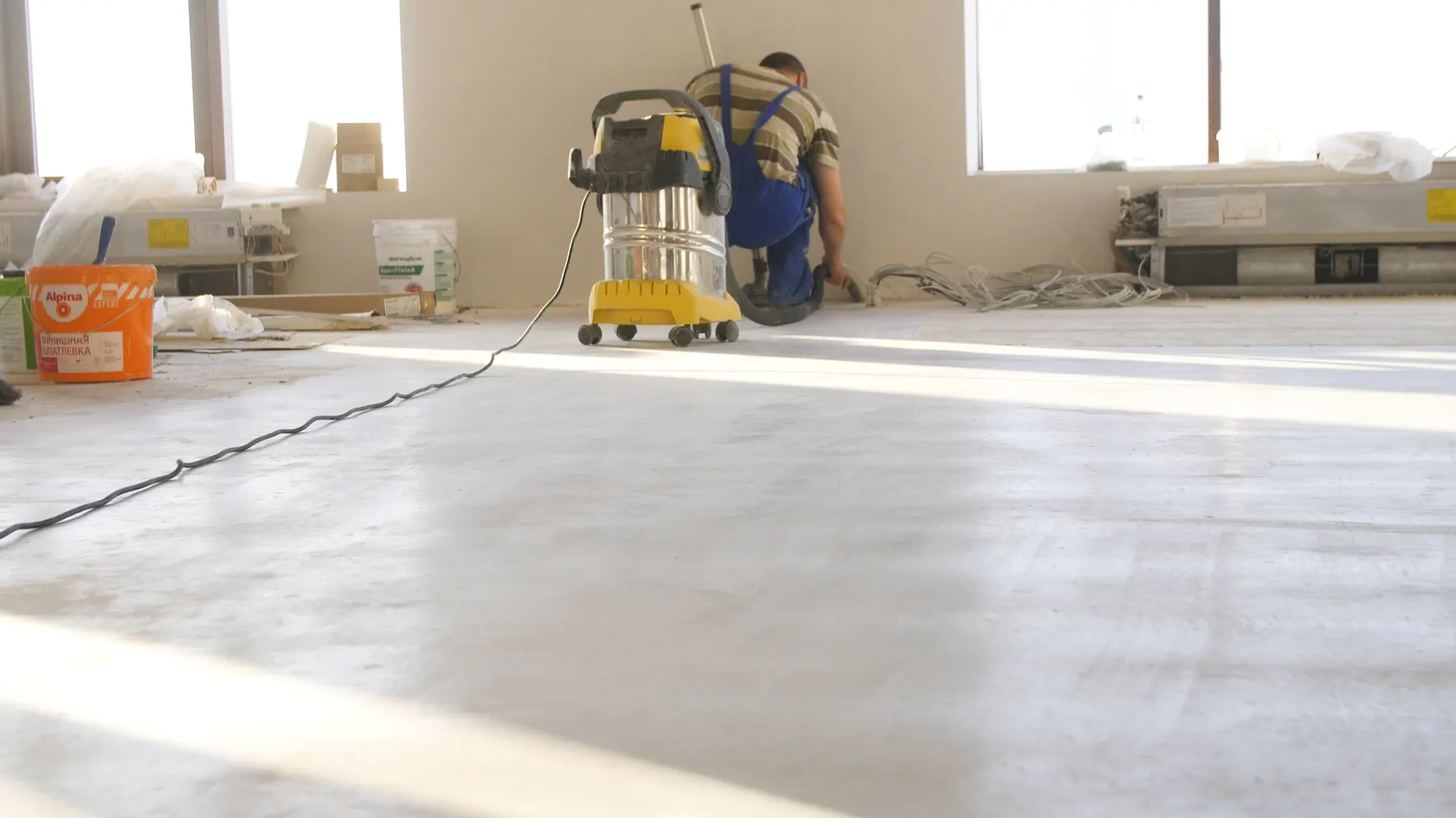A worker in blue overalls vacuuming a bare concrete floor in an unfinished room after building work