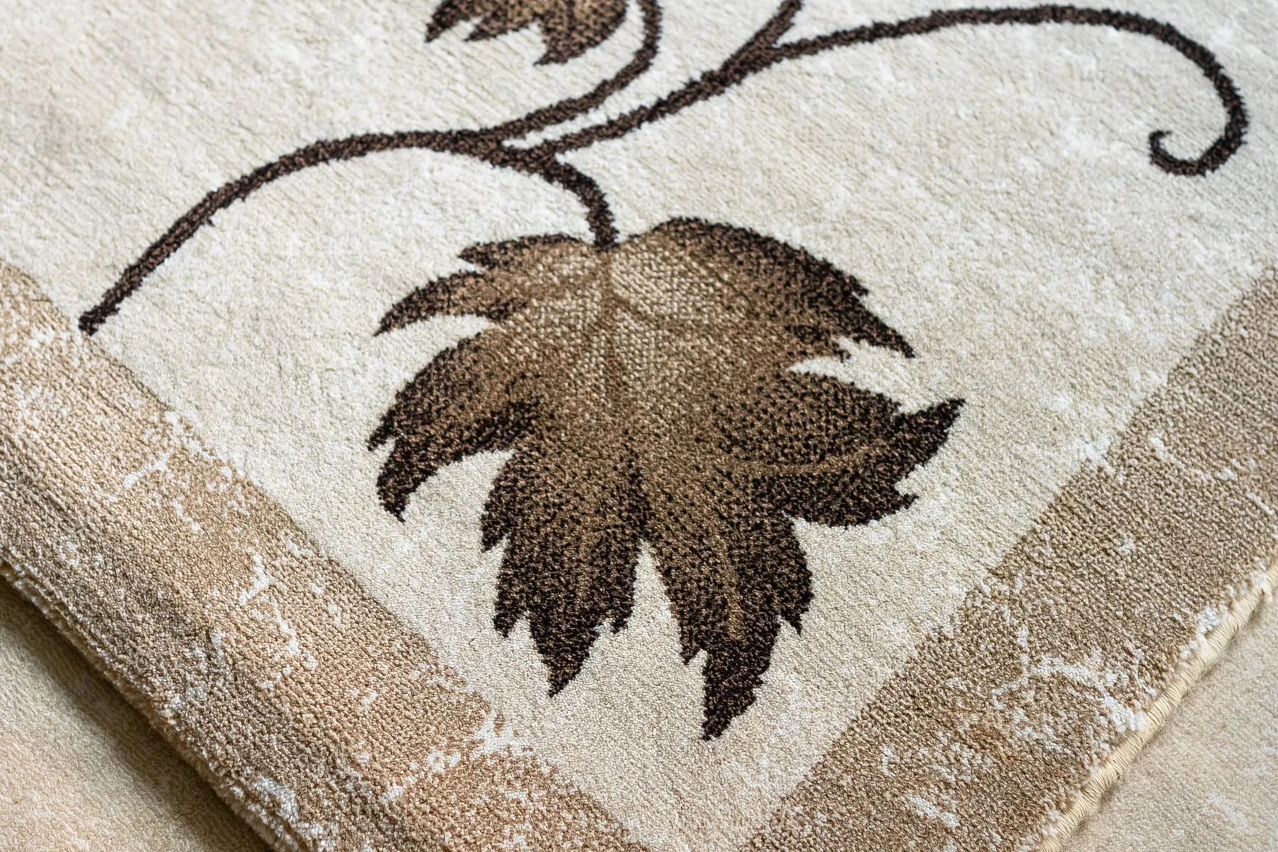 Close-up of a cream area rug with a woven brown leaf and vine motif and a tan border