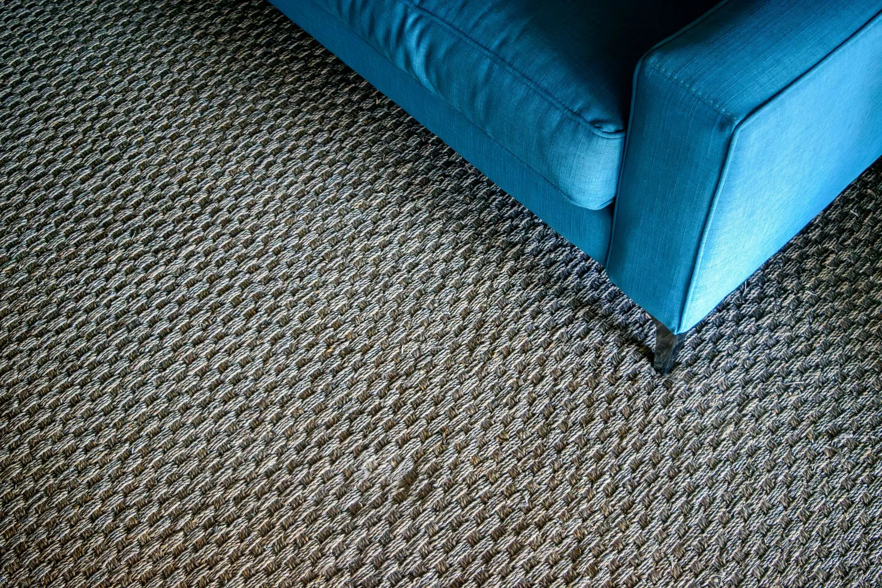 The corner of a teal fabric sofa standing on a dark grey woven loop pile carpet
