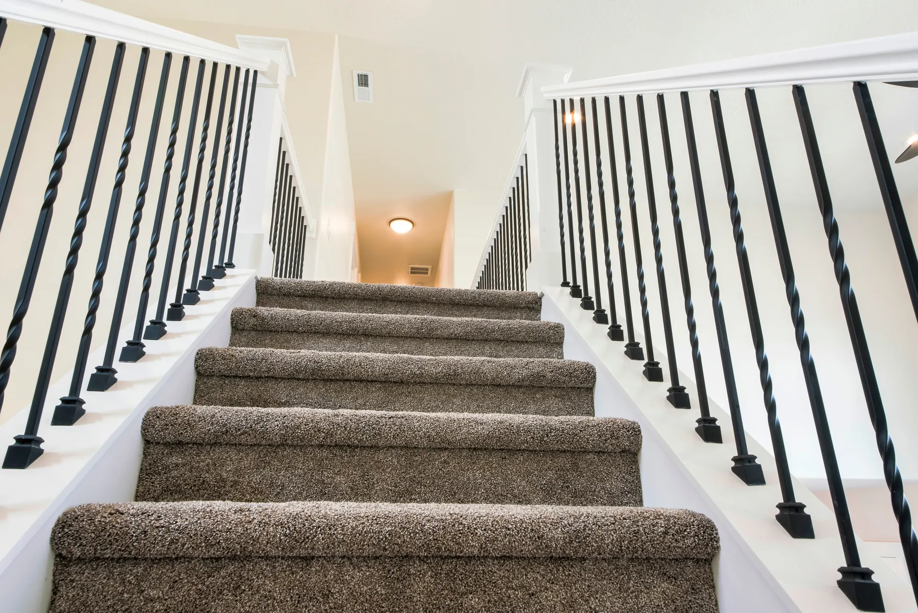 A flight of stairs carpeted in taupe plush pile, between white balustrades with black iron spindles