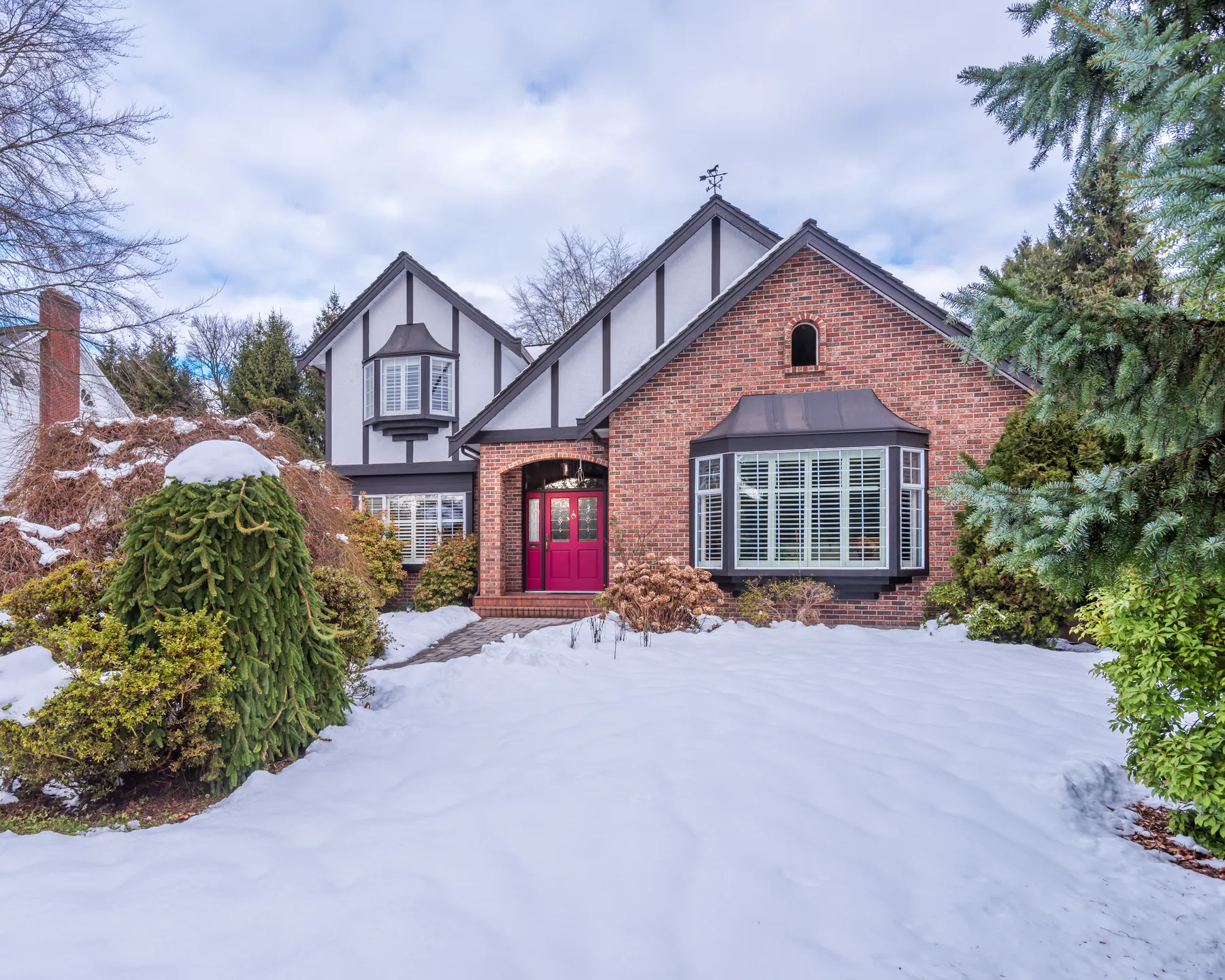 A detached brick and stucco Tudor house behind deep snow, with a red front door and bay windows