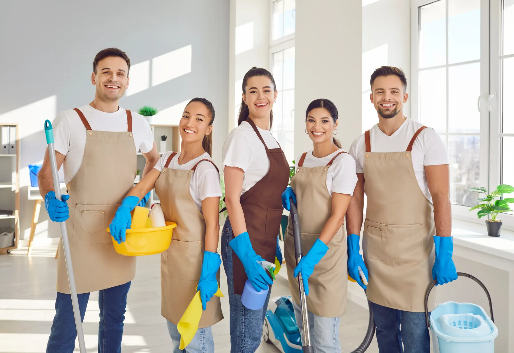Five cleaners in white shirts and beige aprons with mops, gloves and a vacuum in a bright room