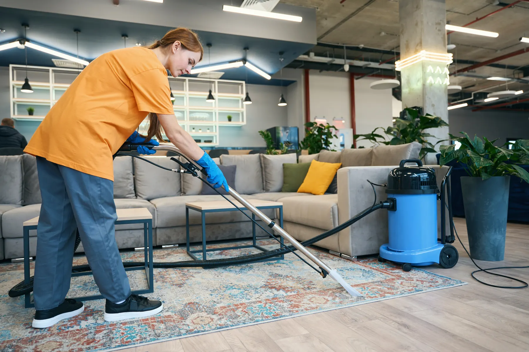 A cleaner in an orange shirt and blue gloves drawing an extraction wand across a patterned rug in an open plan lounge